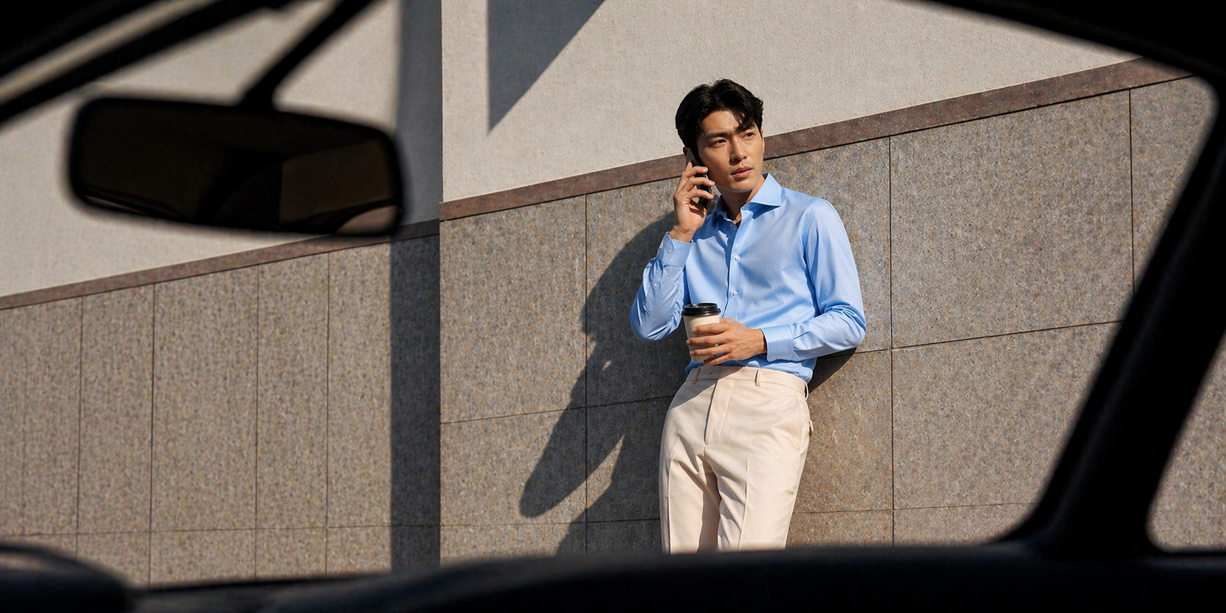Man in a blue shirt and beige pants talking on a phone against a wall.